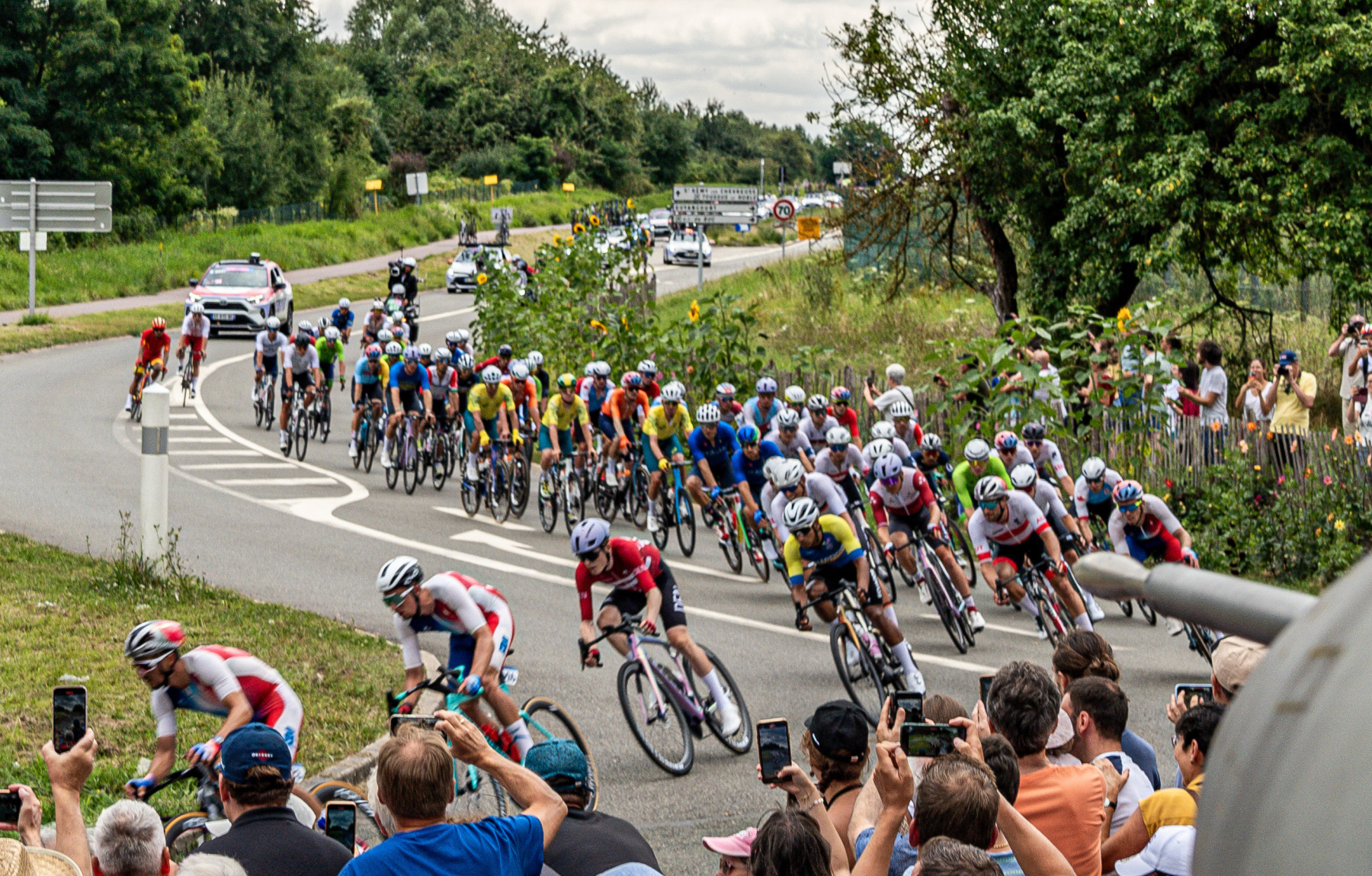 Peloton clustering at the start line with team buses and race officials in the background