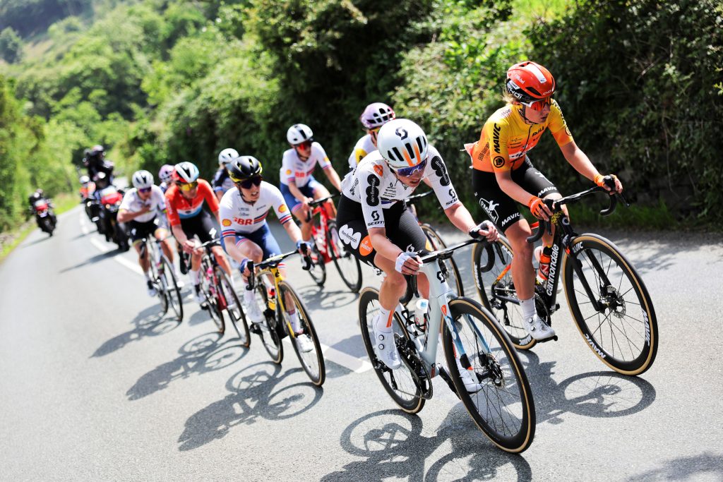 Peloton lining up at the start line under a city skyline with team cars and marshals